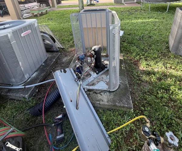 Outdoor condenser with its top and fan removed, exposing the compressor, with refrigerant hoses and tools on the grass around it