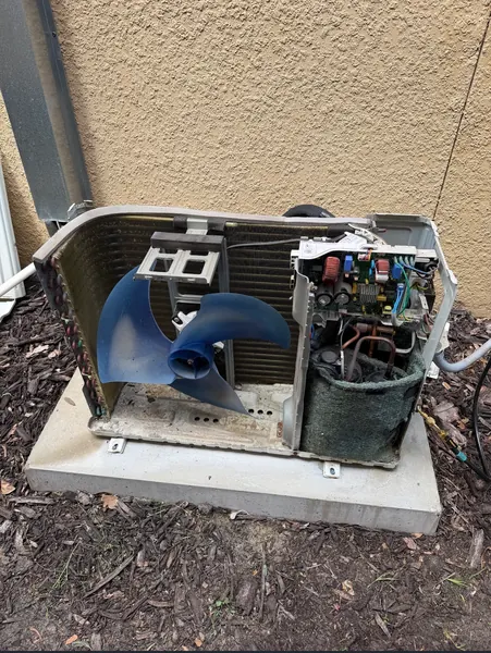 Ductless mini-split outdoor unit with its cover off, showing the blue fan blade, coil and control board, on a pad beside a stucco wall